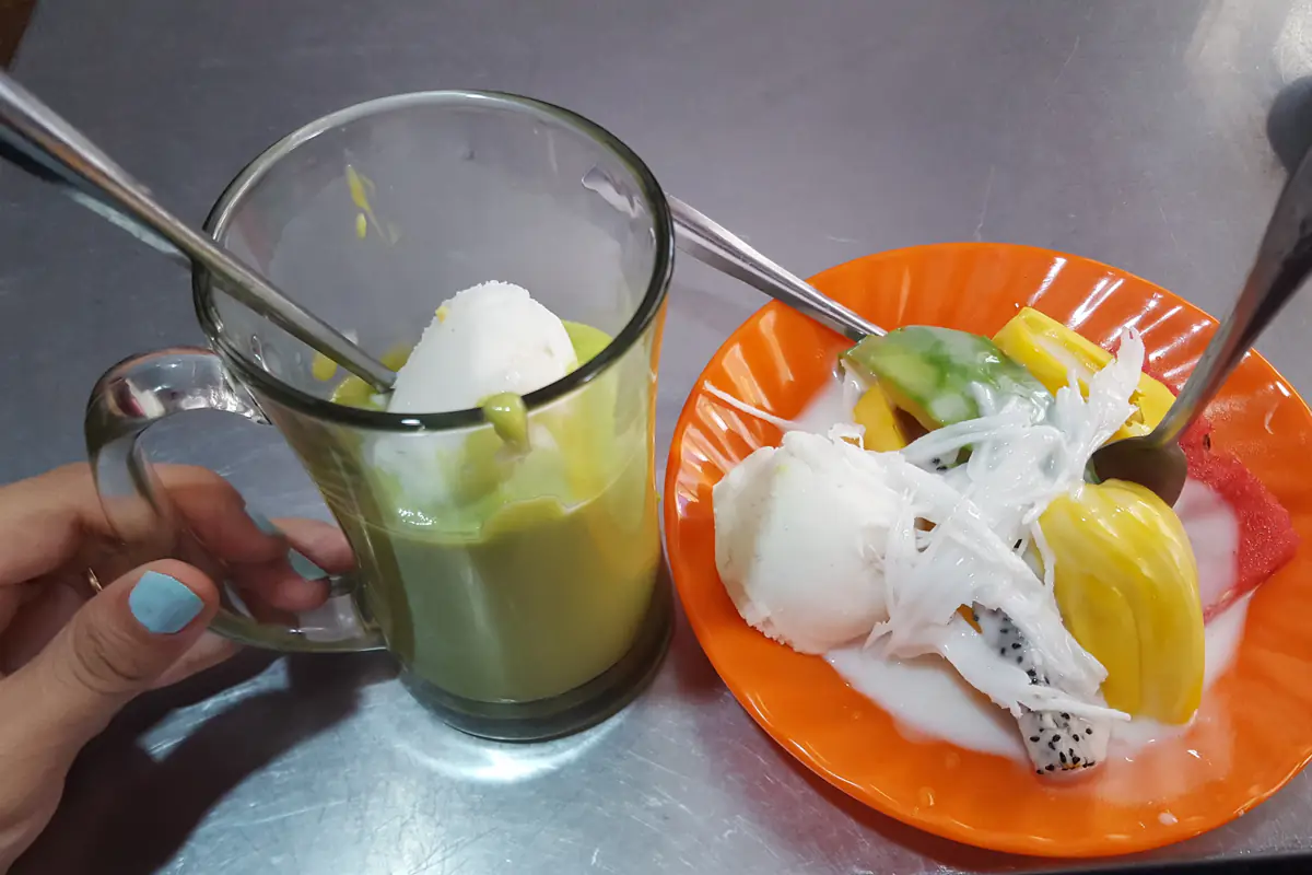Một ly thủy tinh đựng bơ xay màu xanh với viên kem trắng phía trên, đặt cạnh đĩa kem trái cây có mít, thanh long và dừa nạo